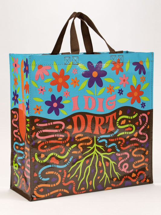 The Blue Q "I Dig Dirt Shopper" is a colorful reusable bag featuring floral and worm illustrations on blue and brown panels, bold “I DIG DIRT” text, and sturdy brown handles.