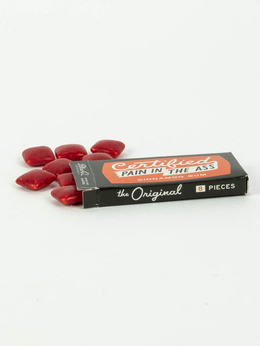 A box of Blue Q's Certified Pain In The Ass Gum, labeled "The Original" and "8 Pieces," features candy-coated gum pieces on a white background—making it a spicy, fun novelty treat for gum lovers.