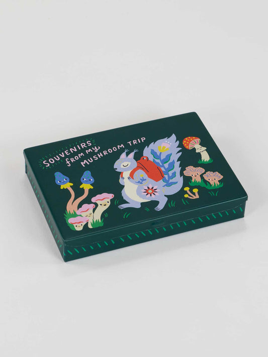 The Blue Q Souvenirs From My Mushroom Trip Pocket Box is a dark green tin decorated with vibrant mushrooms, pebbles, and a squirrel with a red backpack. The lid features “Souvenirs from my mushroom trip” in playful white lettering.