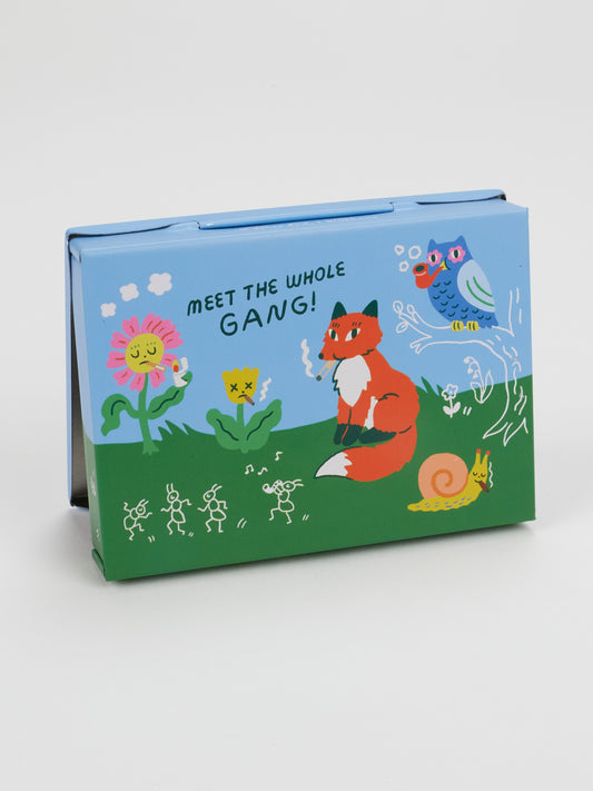 A playful, colorful illustration adorns the Rebel Rabbit Pocket Box by Blue Q, featuring a blue and green box with a flower, two birds, a fox, a rabbit with fuzzy paws, a snail, small stick figures, and the cheerful message “MEET THE WHOLE GANG!”.