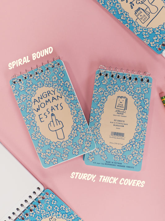 The Blue Q Angry Woman Essays 3x5 Spiral Notebook features a blue floral cover with a cartoon hand flipping the bird. Spiral bound with thick, sturdy covers—perfect as a funny women’s gift or sarcastic decor on any pink desk.