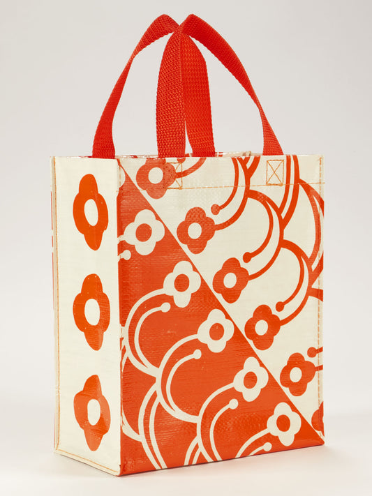 The Blue Q Petal Print Handy Tote features red handles and a bold red and cream geometric floral pattern. This glossy tote stands upright on its own, adding stylish convenience to your daily routine.