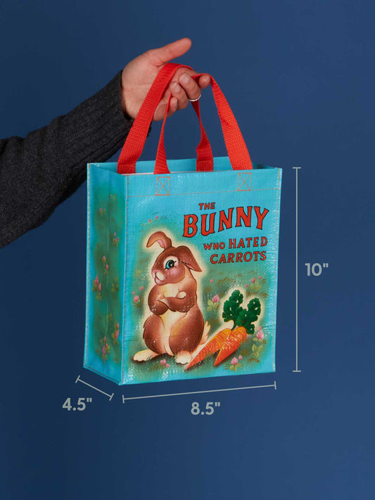 A hand holds Blue Q’s "The Bunny Who Hated Carrots Handy Tote," featuring a bunny, carrots, and red handles. Dimensions: 10" tall, 8.5" wide, 4.5" deep.