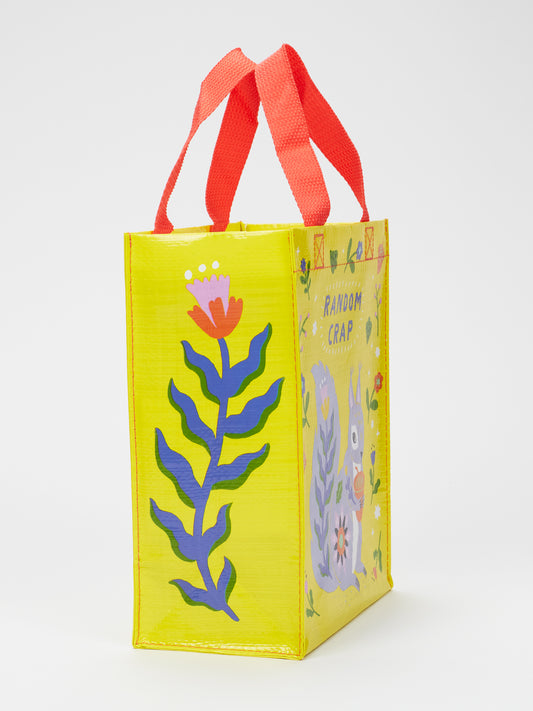 The Blue Q Random Crap Handy Tote is a yellow reusable bag with red handles, made from recycled material, featuring colorful plant and llama illustrations plus "RANDOM CRAP" text, standing upright on a white background.