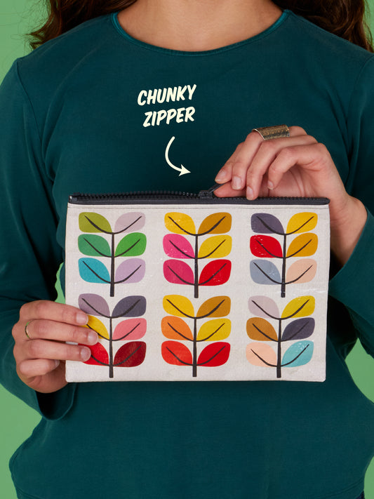 A person wearing a teal shirt and a large ring holds the Blue Q Sprout Zipper Pouch, which features vibrant leaf patterns and a chunky zipper, highlighting its stylish elegance and practical design. The words “CHUNKY ZIPPER” with an arrow emphasize the zipper detail.