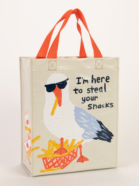 The "I'm Here To Steal Your Snacks Handy Tote" by Blue Q is a reusable bag with orange handles, showing a sunglassed seagull on fries and the text: I’m here to steal your beach snacks.