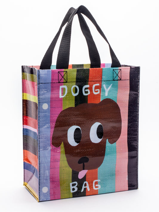 The Blue Q Doggy Handy Tote is a colorful reusable bag with black handles, featuring a cartoon dog face and "DOGGY BAG" text on a striped background.