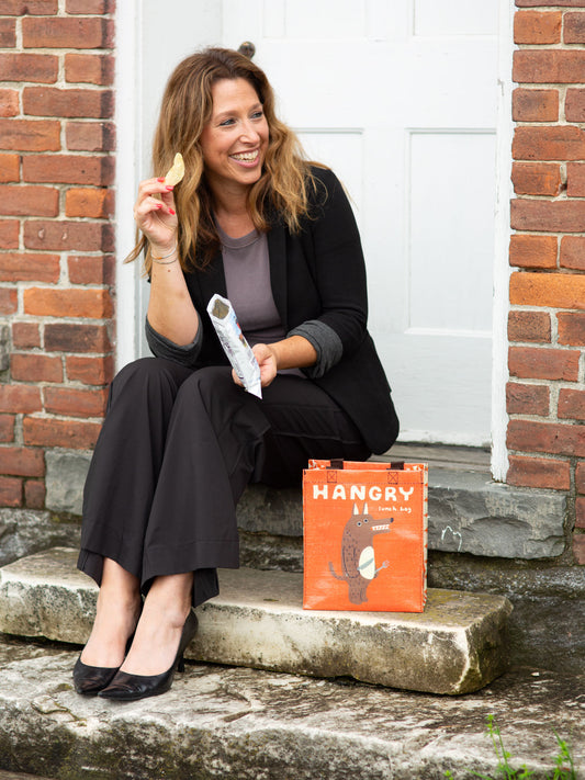 A smiling woman sits on stone steps by a white door, holding a chip and a snack bag. Beside her is the Blue Q Hangry Handy Tote, featuring an illustration of a bear.