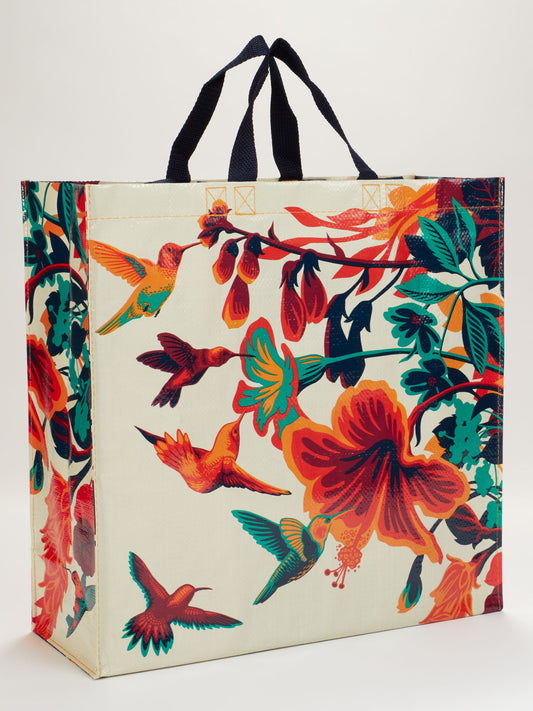 The Blue Q Hummingbird Shopper is a durable reusable bag with black handles, featuring vibrant hummingbirds and bold flowers in red, orange, teal, and green on a light background.