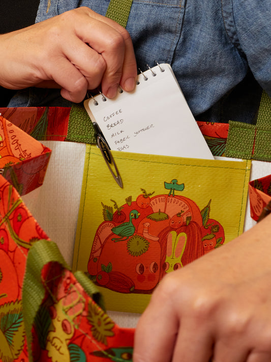 Clutching their handwritten grocery list, a person tucks it into the Blue Q “I’m Living For Tomato Season Shopper” bag, its whimsical animal and fruit design reflecting their excitement to shop for fresh ingredients during peak tomato season.