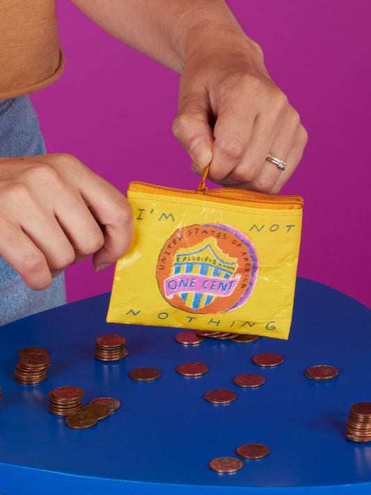 A person holds the Blue Q "I'm Not Nothing Coin Purse," a small yellow pouch with a one-cent design, beside scattered pennies on a blue table, wearing a statement shirt against a pink background.