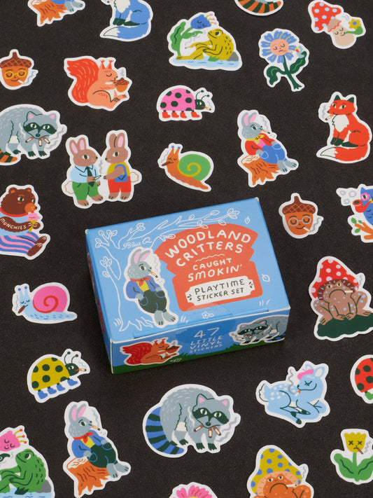 A blue box labeled “Woodland Critters Caught Smokin’ Playtime Sticker Set” by Blue Q is surrounded by colorful stickers featuring cute critters like rabbits, raccoons, foxes, and snails on a black background. Suitable for ages 13 and up.
