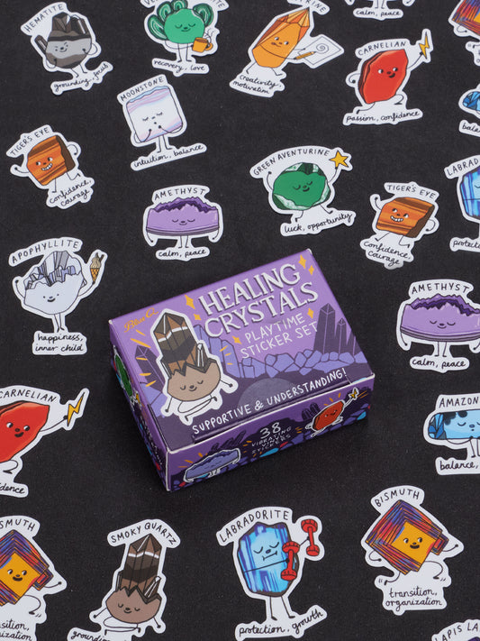 A purple Blue Q Healing Crystals Playtime Sticker Set box rests on a black surface, surrounded by vibrant cartoon crystal stickers—each labeled with the crystal name and a positive trait—making it perfect for inspiring creativity and energy healing in ages 13 and up.