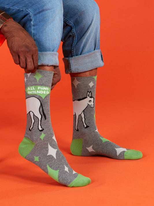 Someone wearing cuffed jeans sits on an orange background, showing Blue Q All Puns Intended M-Crew Socks—gray, with green toes and heels, white stars, a donkey graphic, and “ALL PUNS INTENDED” near the top.