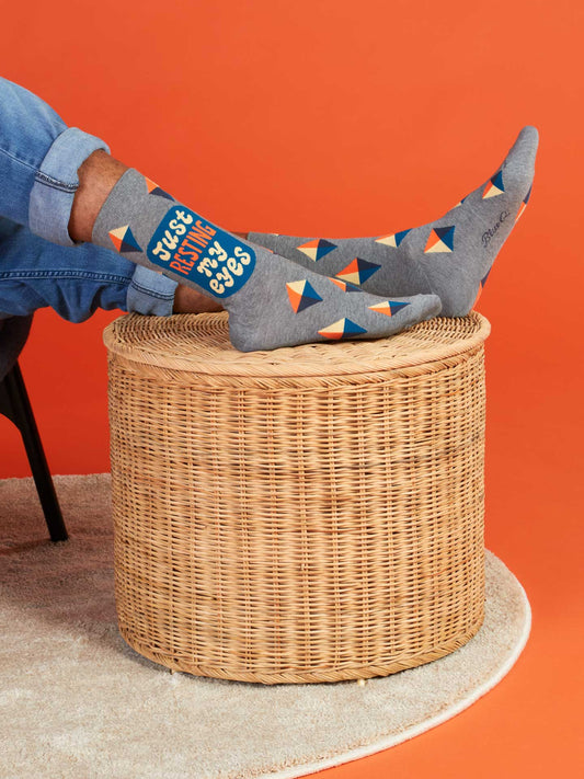 A person lounges with feet up on a round wicker table, wearing Blue Q's Just Resting My Eyes M-Crew Socks—gray combed cotton socks featuring colorful geometric patterns—against an orange background.
