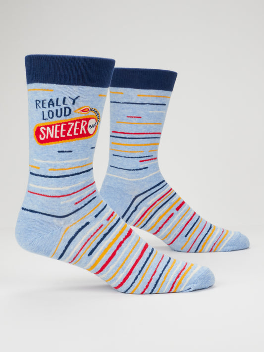 Blue Q’s Really Loud Sneezer M-Crew Socks feature light blue cotton with navy cuffs, toes, and heels, accented by colorful stripes. Designed to fit men’s sizes 7-12, one sock displays the text “REALLY LOUD SNEEZER” next to a small cartoon face holding a tissue.