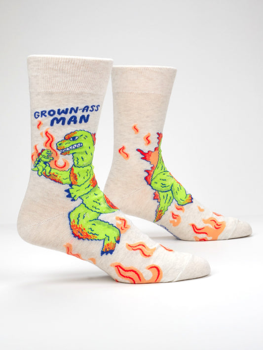 Blue Q’s Grown-Ass Man M-Crew Socks are beige men’s socks showcasing a green cartoon dinosaur holding a drink and surrounded by orange squiggles, with the phrase “GROWN-ASS MAN” above the dinosaur—perfect novelty socks for anyone who loves funny office socks with personality.
