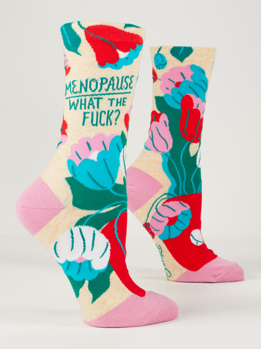 Blue Q’s Menopause. What The Fuck? W-Crew Socks feature abstract floral patterns, bold lettering, and are crafted from combed cotton, nylon, and spandex—vividly showcased on a model’s feet against a plain background.