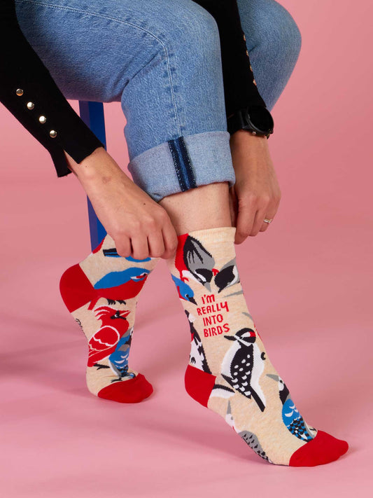 A bird lover sits on a blue stool against a pink background, rolling up their jeans to reveal Blue Q I'm Really Into Birds W-Crew Socks, featuring colorful birds and the phrase "I'M REALLY INTO BIRDS.
