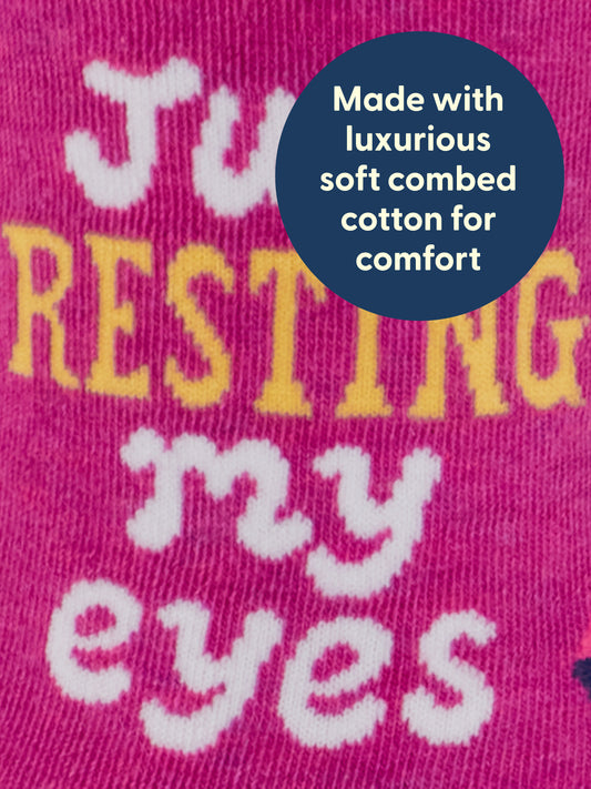 The Blue Q Resting My Eyes W-Crew Socks feature a pink fabric adorned with whimsical yellow and white text that says “Just resting my eyes.” A blue circle highlights that they are made with luxurious, soft combed cotton and a touch of spandex for ultimate comfort. The socks have a soft, stretchy, and textured appearance.