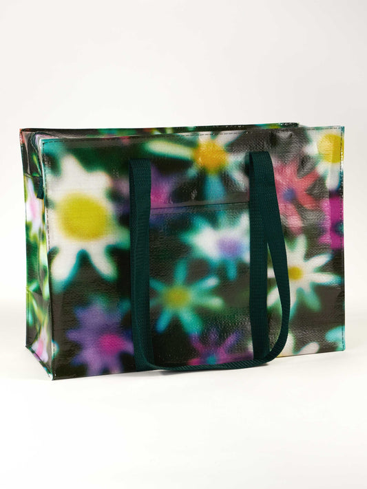 The Blue Q Hazy Daisy Shoulder Tote is a large reusable shopping bag with dark green handles and a vibrant, blurred floral pattern in white, yellow, purple, and green on a glossy finish for a unique look.