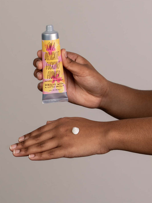 A person applies Blue Q's I'm a Delicate Fucking Flower Natural Hand Cream – Hibiscus with a Little Vanilla onto the back of their hand, a swirl of cream visible on their skin.