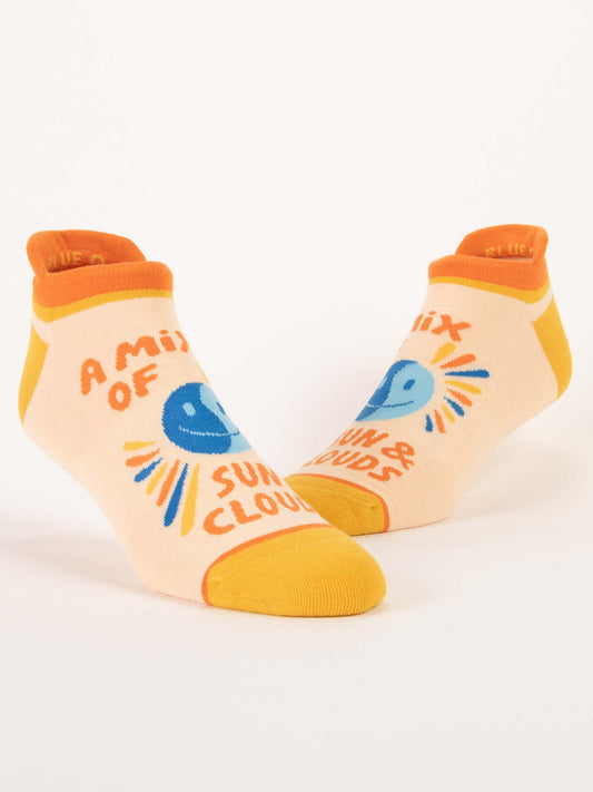 Blue Q’s “A Mix Of Sun & Clouds” Sneaker Socks feature ankle cushioning, orange heels and toes, a blue sun and cloud graphic, and the phrase “A mix of sun & clouds” on a cream background.