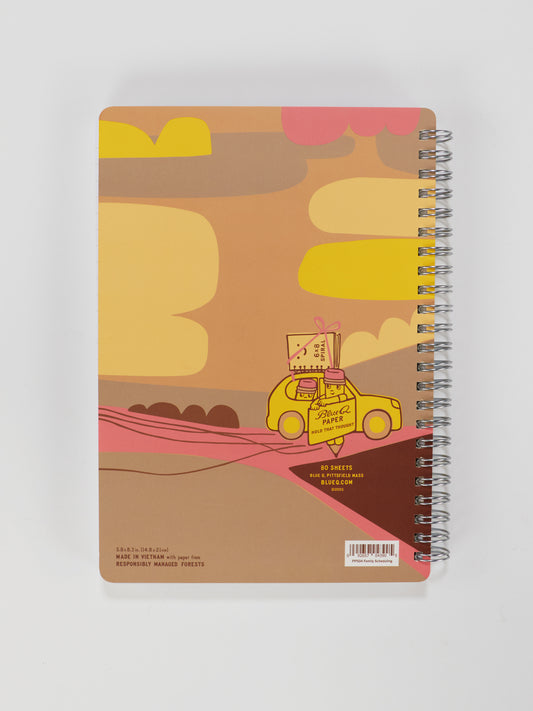 The Blue Q "Adventures In Family Scheduling & Logistics" 6x8 Spiral Notebook features an illustrated cover with a yellow car carrying books and a pencil through a vibrant, whimsical landscape of clouds, hills, and pastel colors.