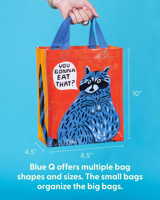 A hand holds the Blue Q "You Gonna Eat That? Handy Tote," featuring a cartoon raccoon and the phrase “You gonna eat that?” Sized 10" tall x 8.5" wide x 4.5" deep, it's a colorful, waste-free bag. Blue Q offers totes in many shapes and sizes.