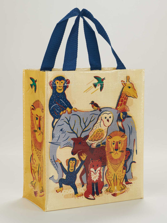 The Blue Q Animal Family Handy Tote is a reusable bag with blue handles, showcasing colorful illustrations of a lion, fox, owl, elephant, chimpanzee, giraffe, and birds on a light yellow background.