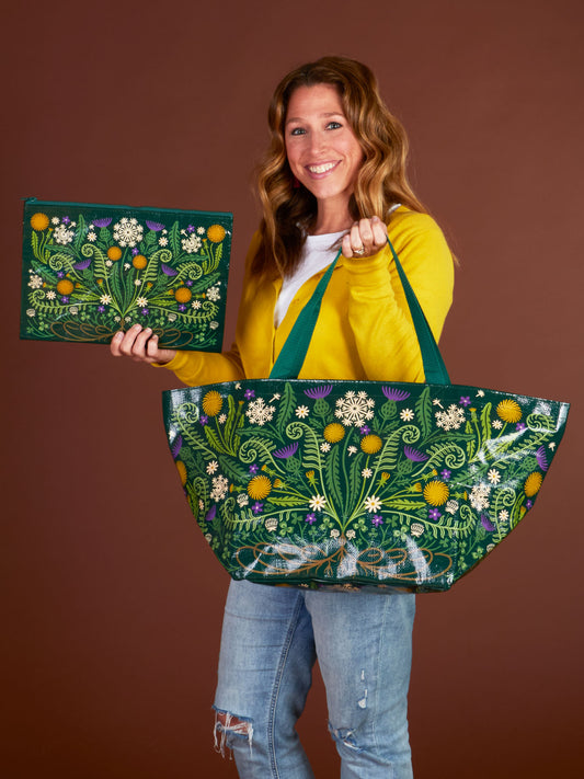 A smiling woman in a yellow cardigan and ripped jeans holds the Blue Q Wild Bloom Big Bag and matching flat pouch, both made from recycled material and adorned with colorful embroidered flowers and leaves, against a brown background.