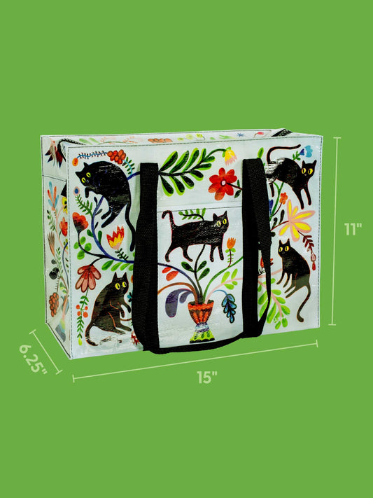 The Blue Q Here Kitty Shoulder Tote is a colorful rectangular bag with black handles, playful black cat and floral designs on white. Sized 15" x 11" x 6.25", it's perfect as a stylish cat carrier, shown against a green background.