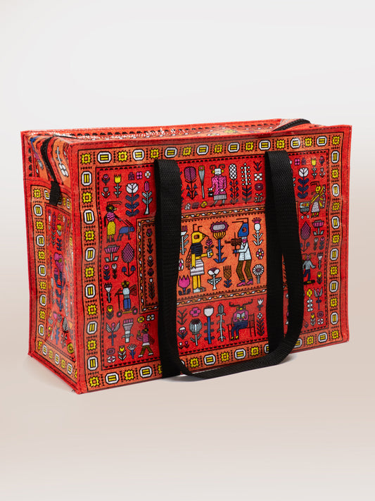 The Blue Q Magic Carpet Shoulder Tote is a sturdy, bright red bag with black handles, made from recycled material. It features folk art people, animals, and plants in geometric frames on a light background and has a secure zipper.