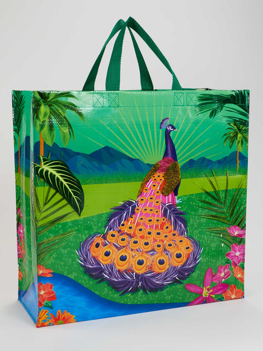The Peacock Paradise Shopper by Blue Q is a reusable tote with green handles, showcasing a vibrant peacock near a blue river with tropical plants and mountains. Made from recycled material, it’s an eco-friendly choice for shopping.