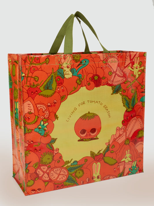 The Blue Q "I'm Living For Tomato Season Shopper" is a colorful reusable tote with green handles, made from recycled materials, featuring whimsical fruits, animals, and a smiling tomato with the words “LIVING FOR TOMATO SEASON.”.