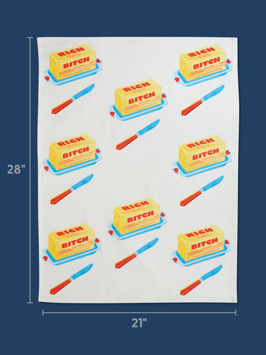 The Blue Q Rich Bitch Dish Towel is a super-absorbent, unbleached cotton kitchen towel (28"x21") featuring a dark blue background with repeating yellow butter sticks labeled "Rich Bitch Butter" and blue-handled red knives.
