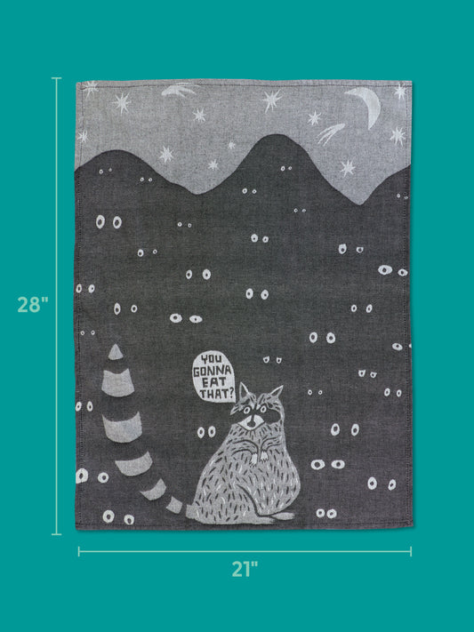 The Blue Q "You Gonna Eat That Dish Towel" (28" x 21") features a cartoon raccoon with a striped tail, glowing eyes, mountains, stars, and a crescent moon on black, asking: “You gonna eat that?”.